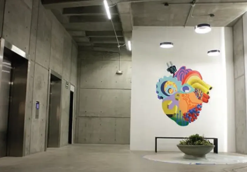 Vestíbulo del edificio MIND en Tijuana: concreto aparente, luminarias circulares y lineales, banca de herrería y mural
